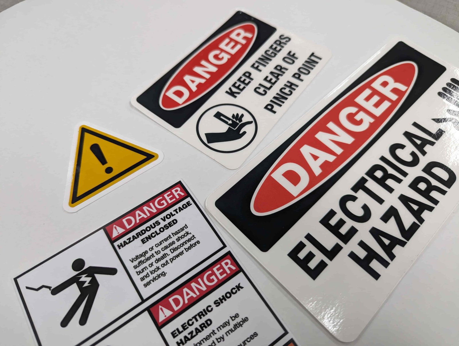 Four custom machine decals on a white background arranged in two rows of two. In the upper left is a small yellow triangle with an exclamation mark on it. The other three are all white rectangles with black text that give warnings about electrical hazards and keeping fingers clear of machinery.